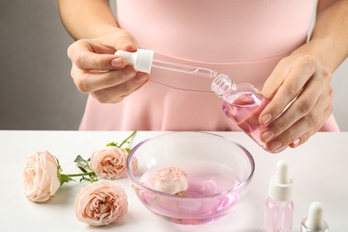 Young woman dripping rose essential oil into glass bottle at table, closeup