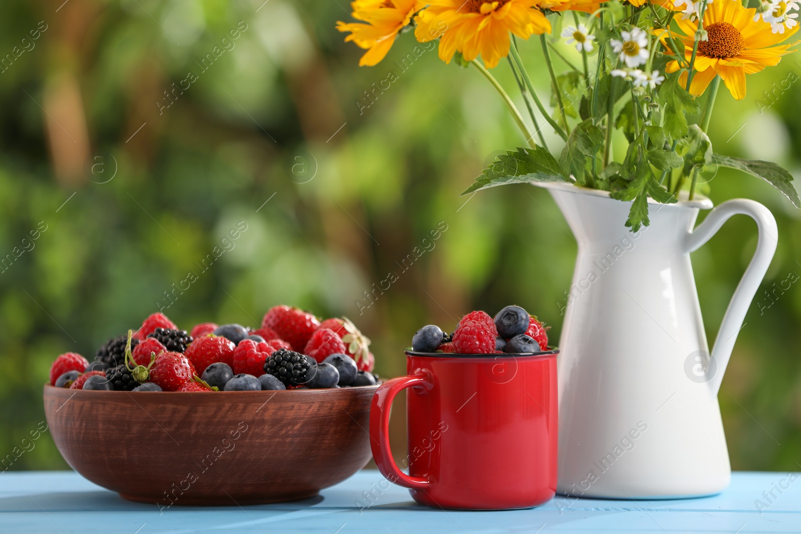 Photo of Different fresh ripe berries and beautiful flowers on light blue table outdoors