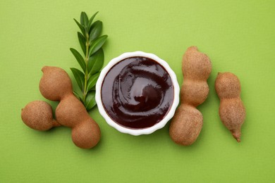 Photo of Bowl of tasty tamarind sauce, leaves and fresh pods on light green table, flat lay
