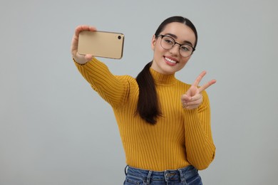 Photo of Smiling young woman taking selfie with smartphone and showing peace sign on grey background