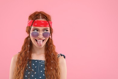 Photo of Stylish young hippie woman in sunglasses showing her tongue on pink background, space for text