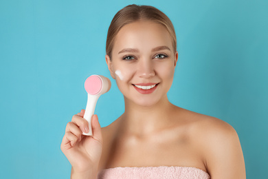 Photo of Young woman washing face with brush and cleansing foam on light blue background. Cosmetic products