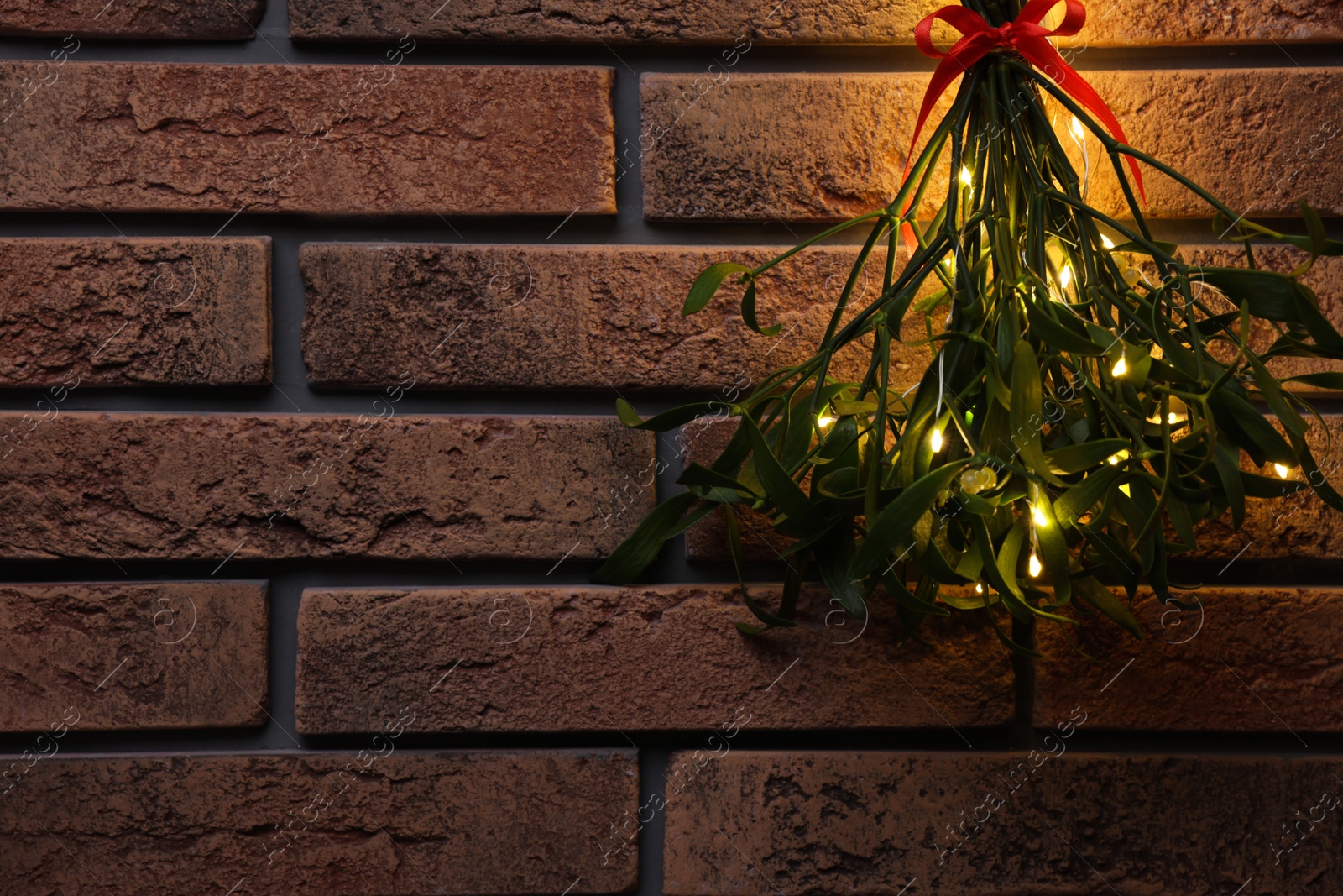 Photo of Mistletoe bunch with red bow and fairy lights hanging on brick wall, space for text. Traditional Christmas decor