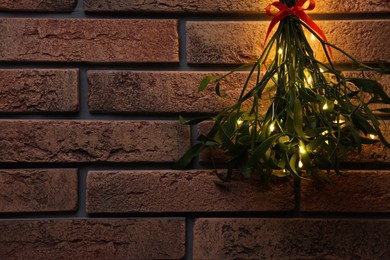 Photo of Mistletoe bunch with red bow and fairy lights hanging on brick wall, space for text. Traditional Christmas decor