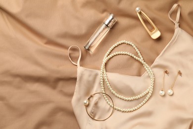 Stylish jewelry with pearls, luxury perfume and silk dress on beige fabric, flat lay