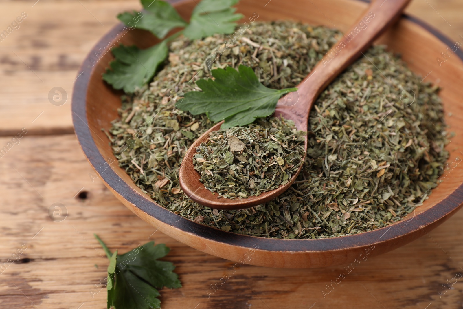 Photo of Bowl and spoon with dried parsley on wooden table, closeup