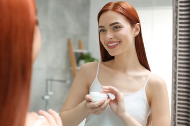 Photo of Beautiful young woman holding jar of body cream near mirror in bathroom