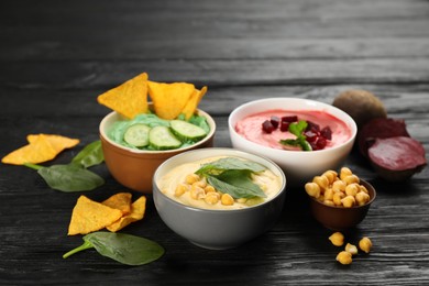 Photo of Different kinds of tasty hummus, nachos and ingredients on black wooden table