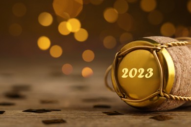 Image of Cork of sparkling wine and muselet cap with engraving 2023 on wooden table against blurred festive lights, closeup. Bokeh effect