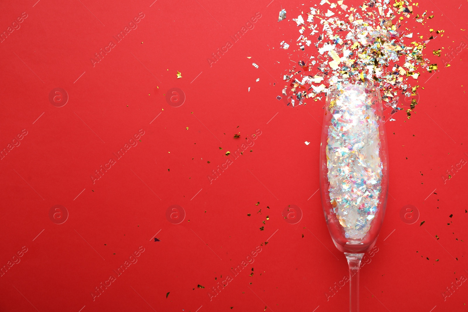 Photo of Shiny confetti spilled out of champagne glass on red background, top view. Space for text