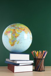 Globe, school supplies and books on wooden table near green chalkboard. Geography lesson