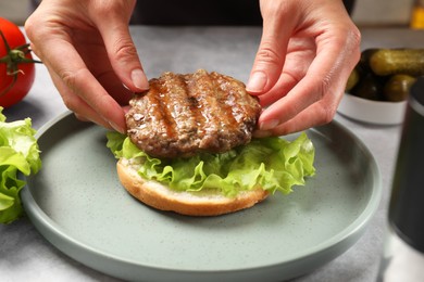 Photo of Woman making tasty hamburger with fried patty, lettuce and bun at table, closeup