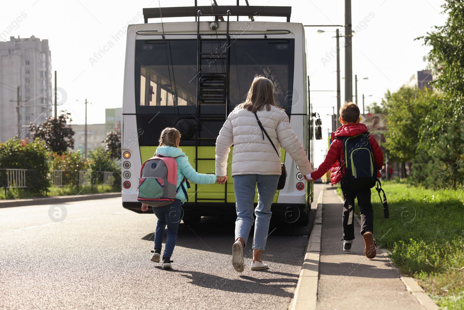 Photo of Being late for school. Senior woman and her grandchildren with backpacks running towards bus outdoors, back view