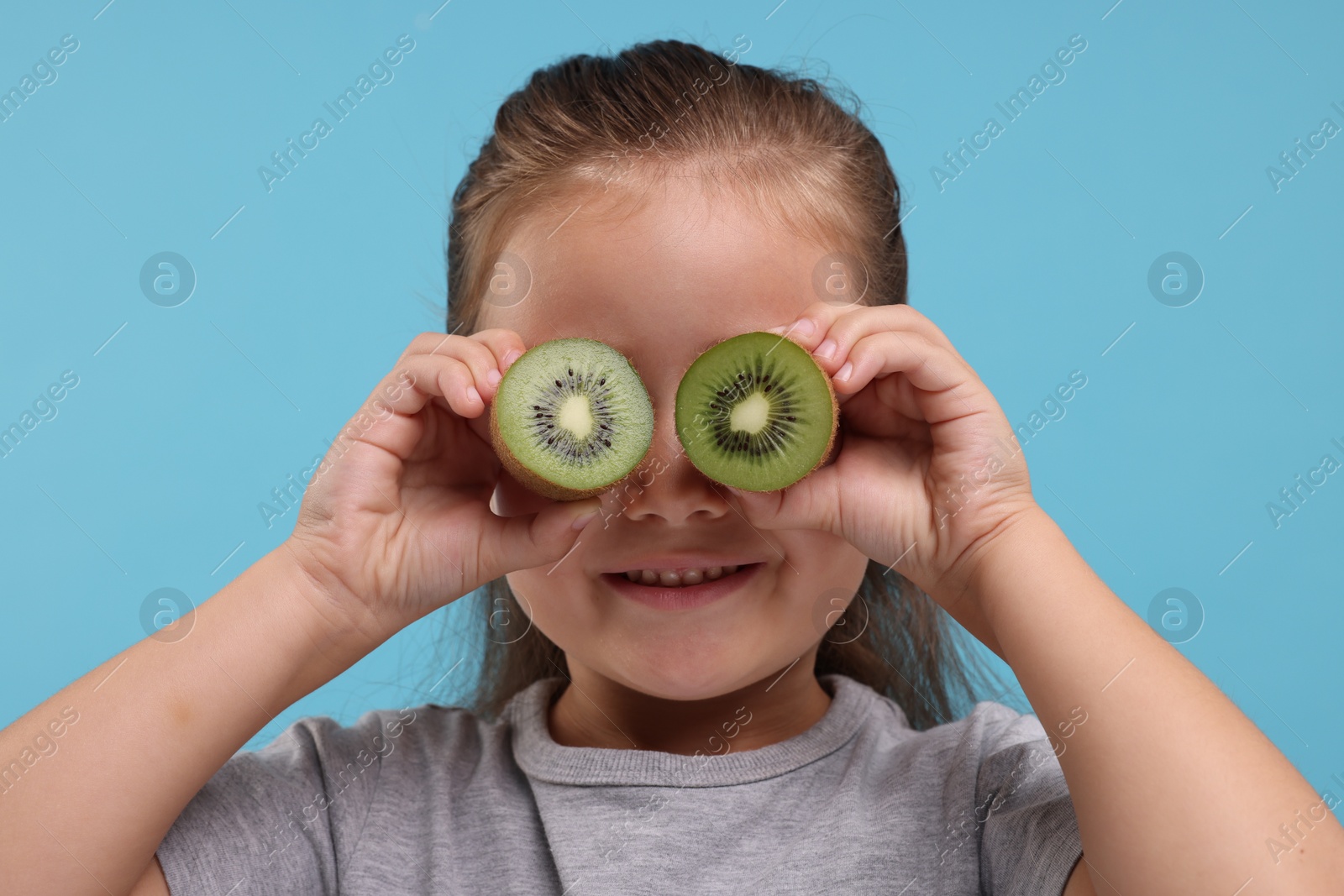 Photo of Smiling girl covering eyes with halves of fresh kiwi on light blue background