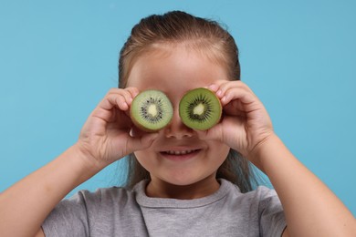 Smiling girl covering eyes with halves of fresh kiwi on light blue background