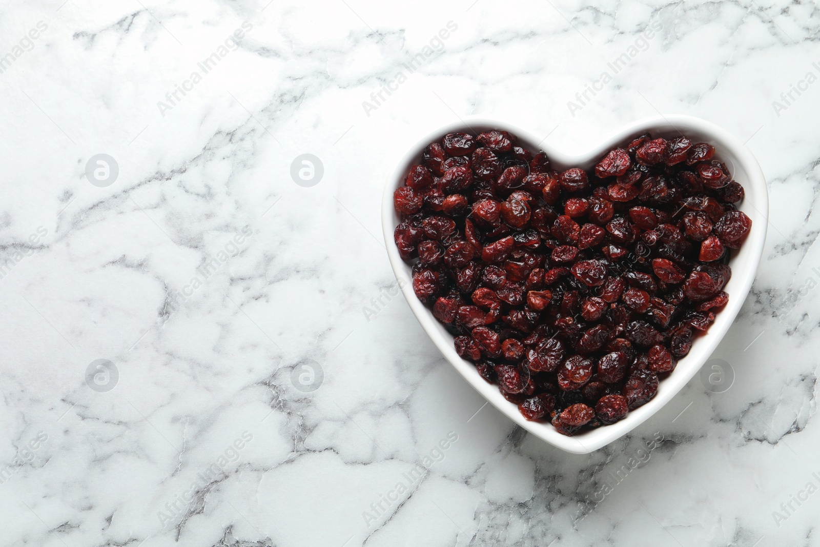 Photo of Heart shaped bowl with cranberries on marble table, top view with space for text. Dried fruit as healthy snack
