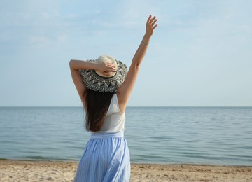 Beautiful young woman wearing straw hat on beach, back view. Stylish headdress