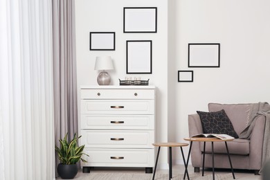 Photo of Empty frames hanging on white wall, chest of drawers and wooden tables indoors