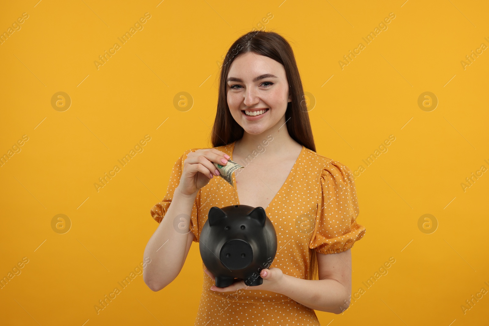 Photo of Happy woman putting dollar banknote into piggy bank on orange background