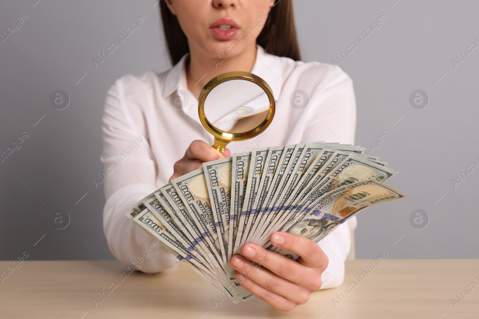 Photo of Expert authenticating 100 dollar banknotes with magnifying glass at table on light grey background, closeup. Fake money concept
