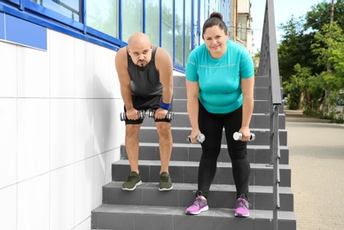 Photo of Tired overweight man and woman with dumbbells on stairs outdoors
