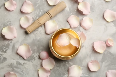 Golden singing bowl with petals and mallet on grey table, flat lay. Sound healing