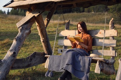 Photo of Beautiful young woman reading book on wooden swing outdoors