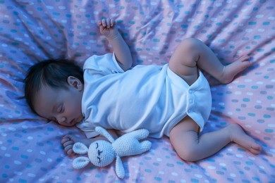 Photo of Cute newborn baby sleeping with toy bunny in crib at night, top view