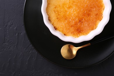 Photo of Delicious creme brulee in bowl served on dark gray textured table, top view