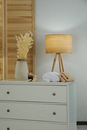 Vase with dried flowers, stacked towels and candles on chest of drawers in room. Interior design
