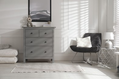 Stylish room interior with grey chest of drawers and comfortable chair