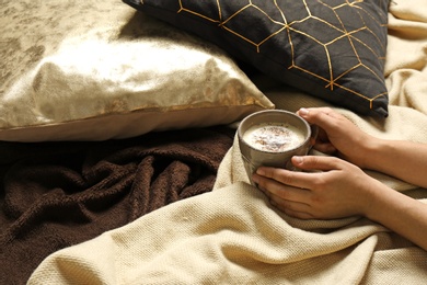 Woman holding cup of coffee while lying on bed with pillows and warm plaid, closeup