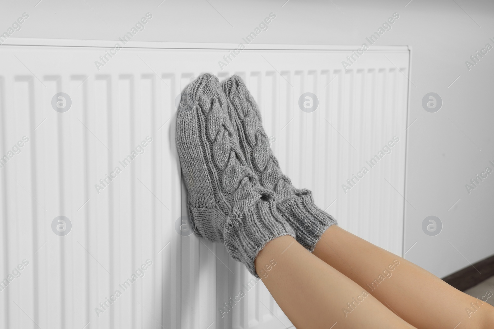 Photo of Woman warming legs on heating radiator near white wall, closeup