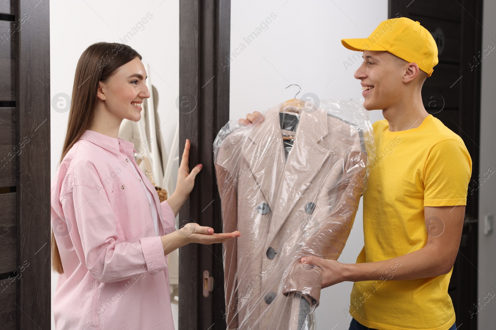 Photo of Dry-cleaning delivery. Courier giving coat in plastic bag to woman indoors