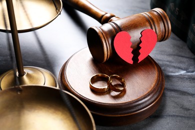 Photo of Divorce concept. Wooden gavel with wedding rings and broken red paper heart on black table, closeup