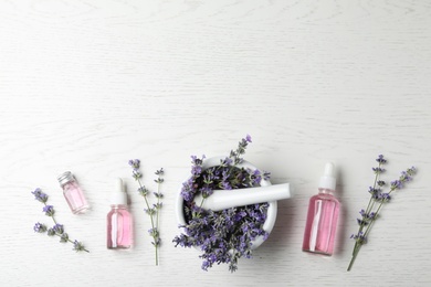 Bottles of essential oil, mortar and pestle with lavender flowers on white wooden background, flat lay. Space for text