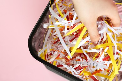 Photo of Woman putting shredded colorful paper strips into trash bin on pink background, closeup