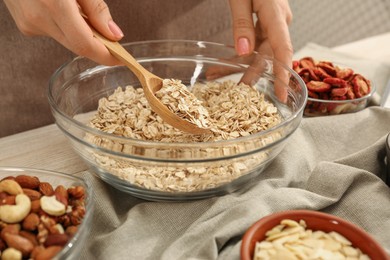 Photo of Making granola. Woman putting oat flakes into bowl at table in kitchen, closeup