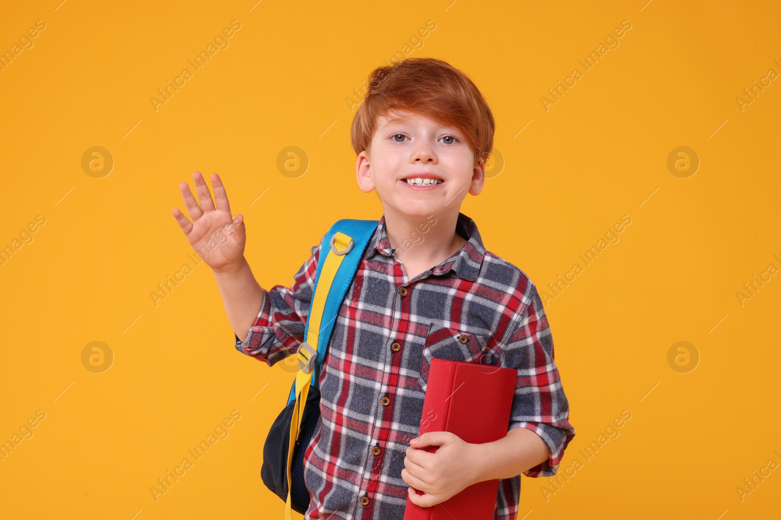 Photo of Happy schoolboy with backpack and book waving hello on orange background