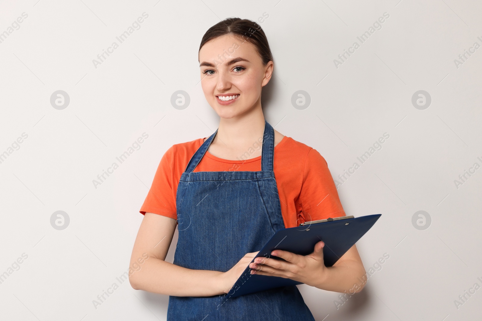 Photo of Beautiful young woman in clean denim apron with clipboard on light grey background