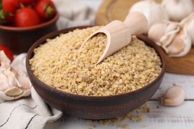 Photo of Bowl and scoop with raw bulgur on white wooden table, closeup