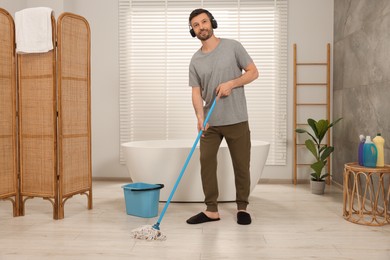 Photo of Enjoying cleaning. Man in headphones listening to music and mopping floor in bathroom