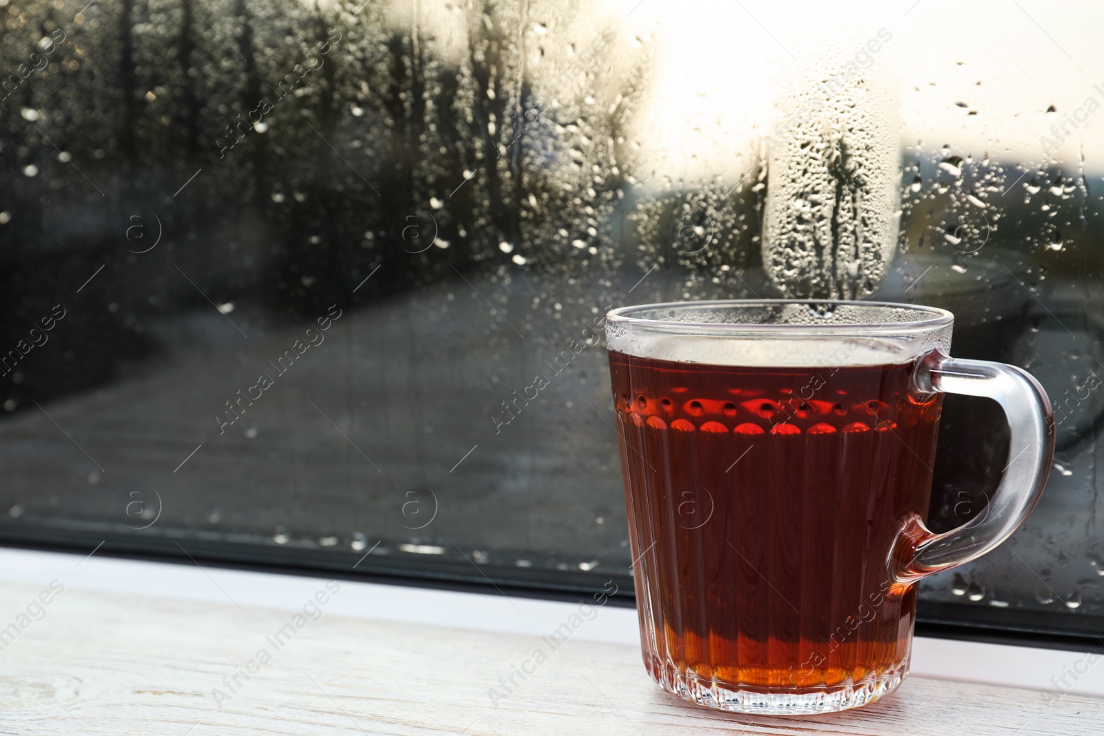 Photo of Glass mug of hot tea on white wooden window sill, closeup. Rainy weather