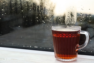 Photo of Glass mug of hot tea on white wooden window sill, closeup. Rainy weather