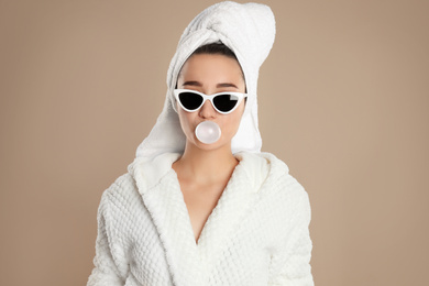 Photo of Young woman in bathrobe and sunglasses blowing chewing gum on brown background