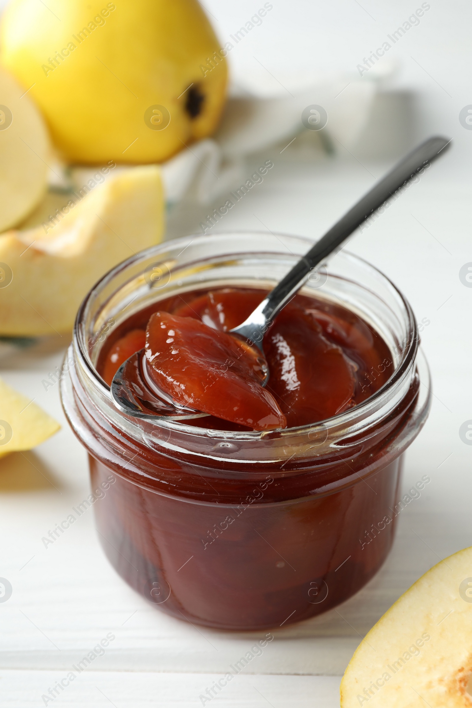 Photo of Tasty homemade quince jam in jar, spoon and fruits on white wooden table, closeup