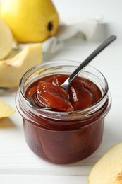 Tasty homemade quince jam in jar, spoon and fruits on white wooden table, closeup
