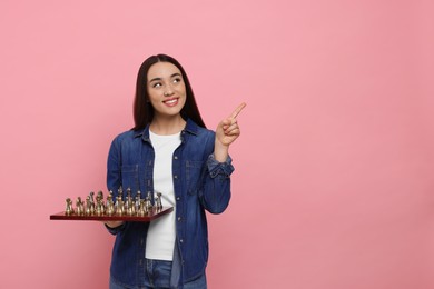 Happy woman with chessboard pointing upwards on pink background, space for text