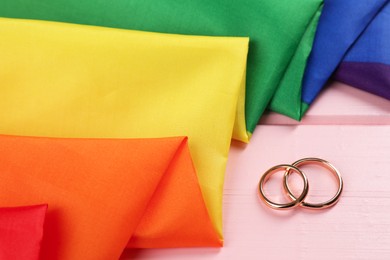 Rainbow LGBT flag and wedding rings on pink wooden table, closeup