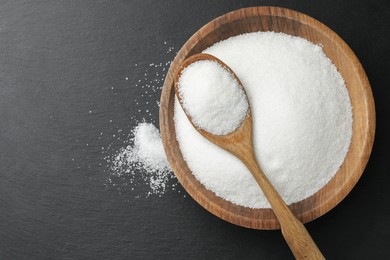 Granulated sugar in bowl and spoon on black table, top view. Space for text